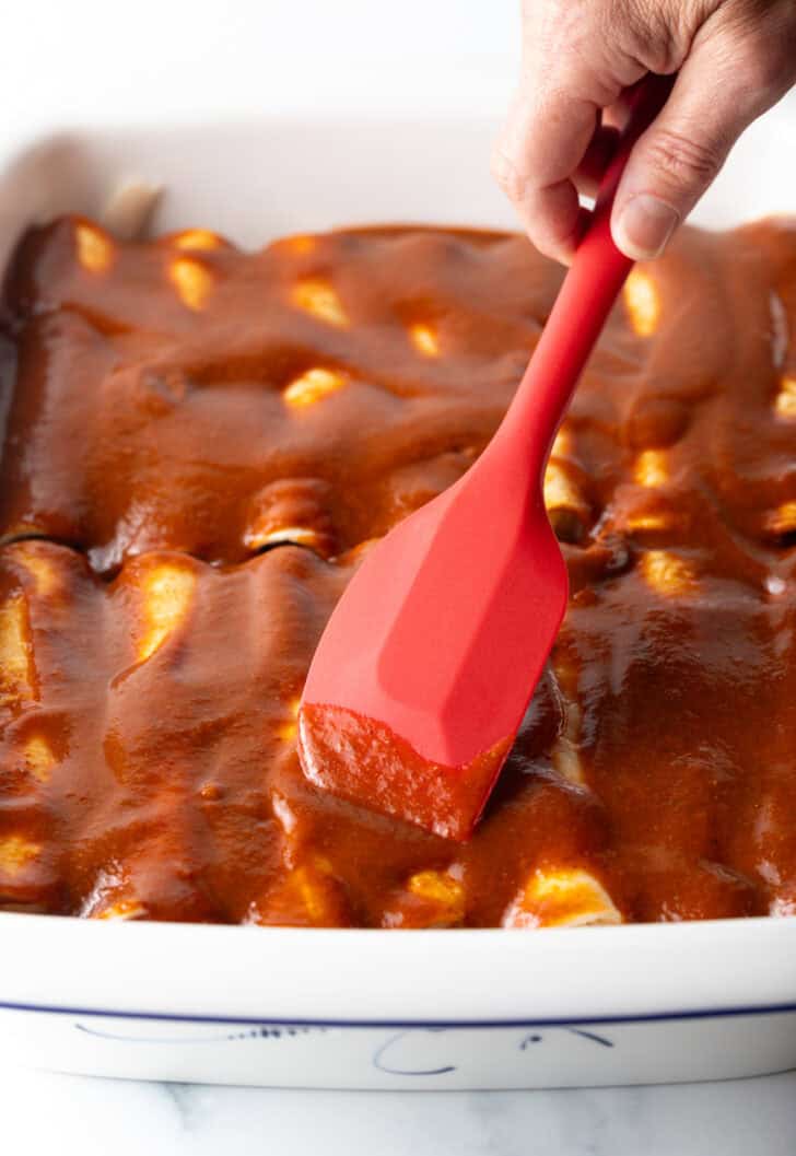 Using a red spatula to smooth red sauce over the unbaked rolled enchiladas.