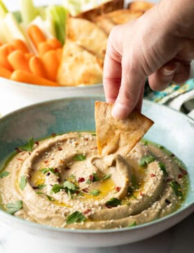 Hand using a pita chip to scoop eggplant dip from a light blue bowl.