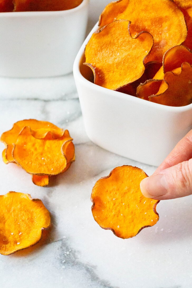 Hand holding a sweet potato chip towards the camera with more chips in the background.