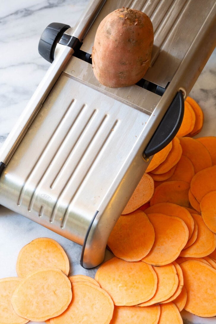 Sweet potatoes being sliced on a mandolin slicer.