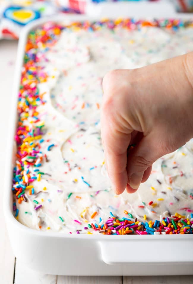 Hand sprinkling colorful sprinkles around the edges of the iced cake.