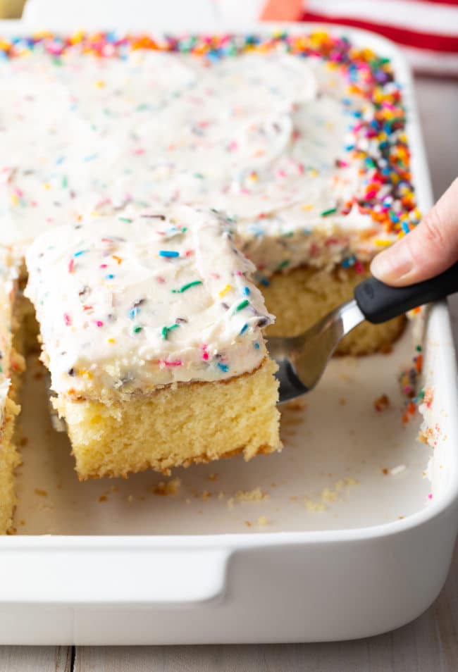 Hand holding a spatula to lift a piece of cake out of the baking dish full of vanilla cake.