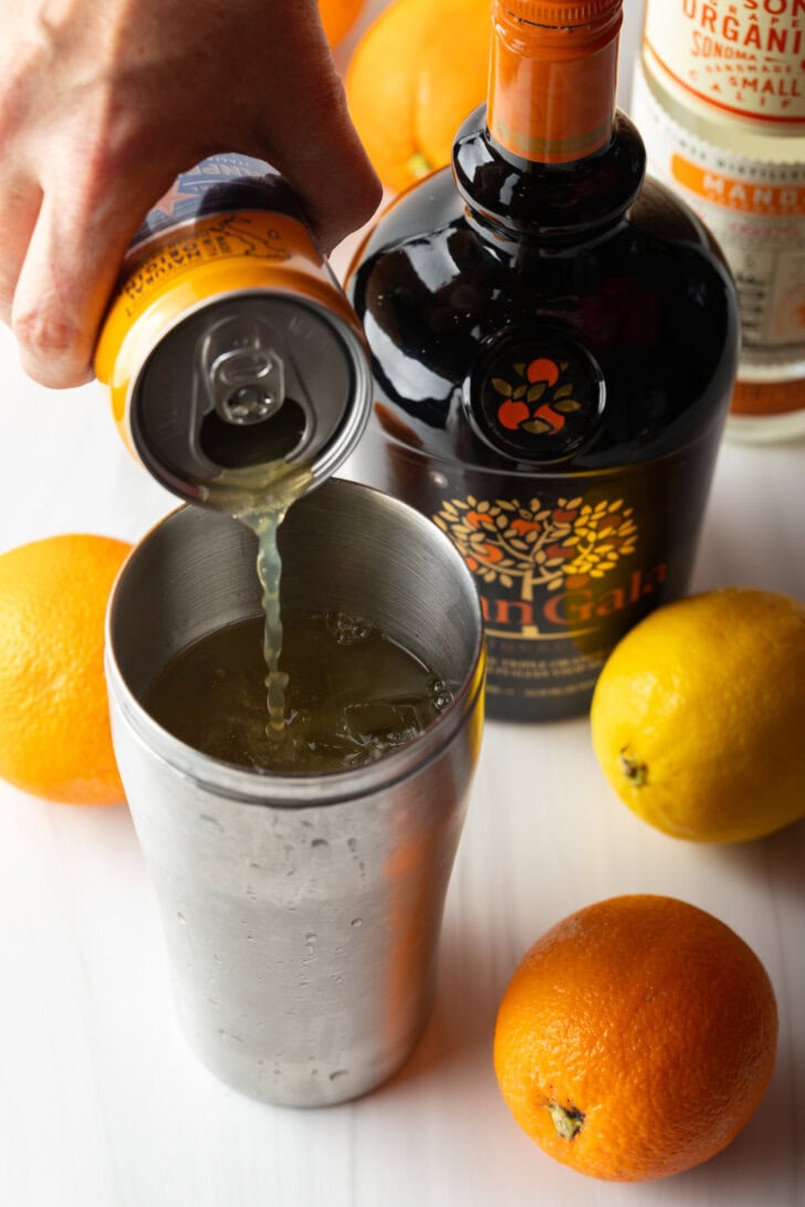 Hand pouring orange soda into metal cocktail shaker.