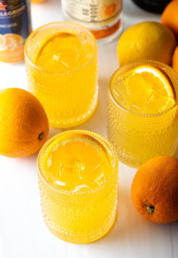 Three orange crush cocktails in glasses, each garnished with a slice of orange. There are fresh oranges and lemons around the glasses.