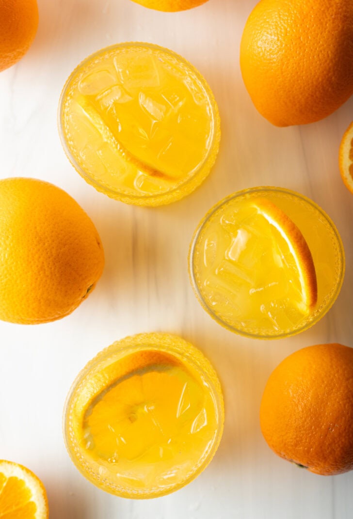 Top down view 4 orange crush drinks in glasses, each garnished with a slice of orange. There are fresh oranges and lemons around the glasses.