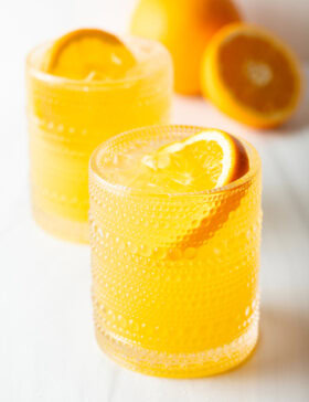 Two orange crush cocktails in glasses, each garnished with a slice of orange. There are fresh oranges and lemons around the glasses.