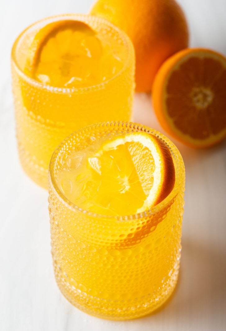 Two orange crush cocktails in glasses, each garnished with a slice of orange. There are fresh oranges and lemons around the glasses.