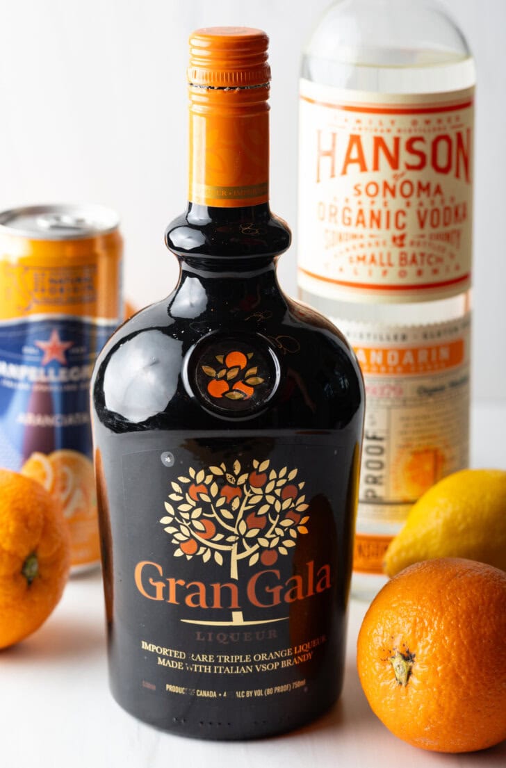 Recipe ingredients: Bottle of Grangala in the front, fresh oranges, bottle of vodka, and can of orange soda in the background.