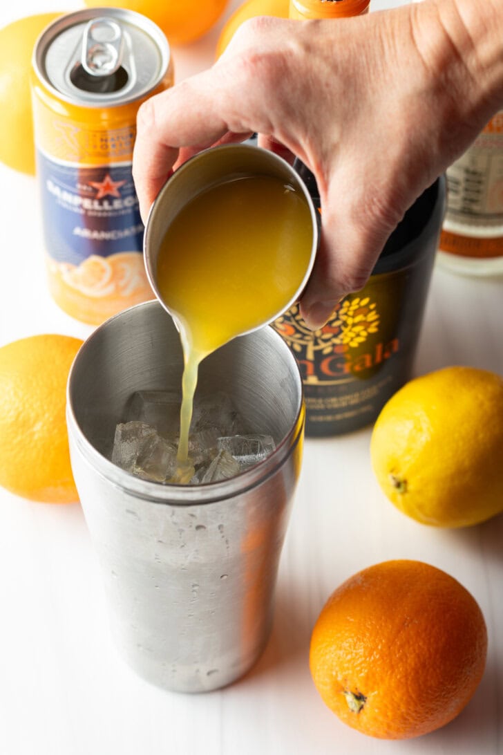 Hand adding orange juice from metal glass into a metal shaker.
