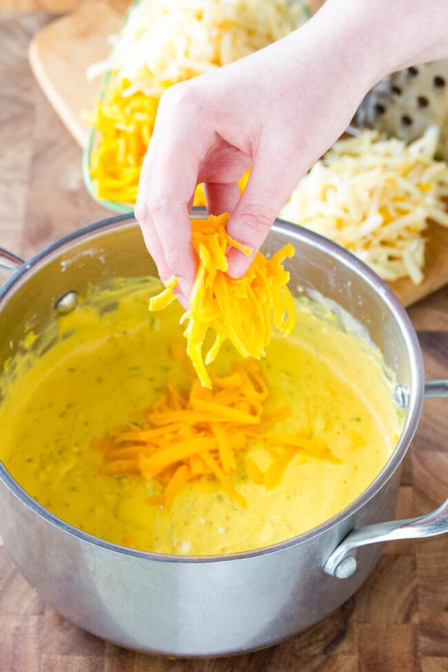 Queso sauce makes or breaks any nacho recipe. The Best Queso recipe is easy and flavorful, with sharp and smoky notes. Start dipping those tortilla chips! #ASpicyPerspective #queso #cheese #dip Dropping shredded cheese in pot.
