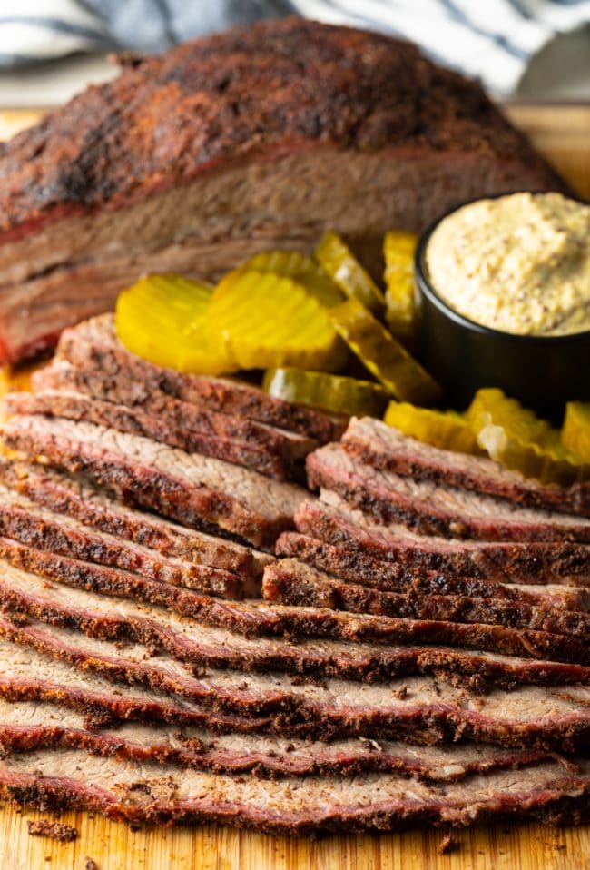 Smoked beef brisket sliced on a wooden cutting board and served with pickles and mustard.