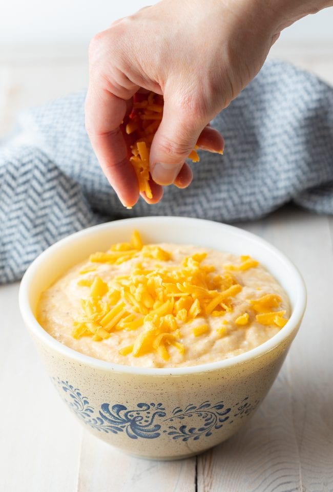 Bowl of grits with a hand above it sprinkling cheese on top.