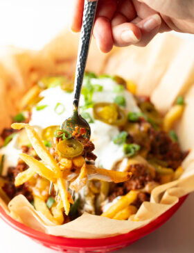 Basket with parchment paper, filled with crispy fries topped with chili and melted cheddar cheese, sour cream, chopped green onions, and jalapeno slices. A hand with forkful of fries is showing to camera.
