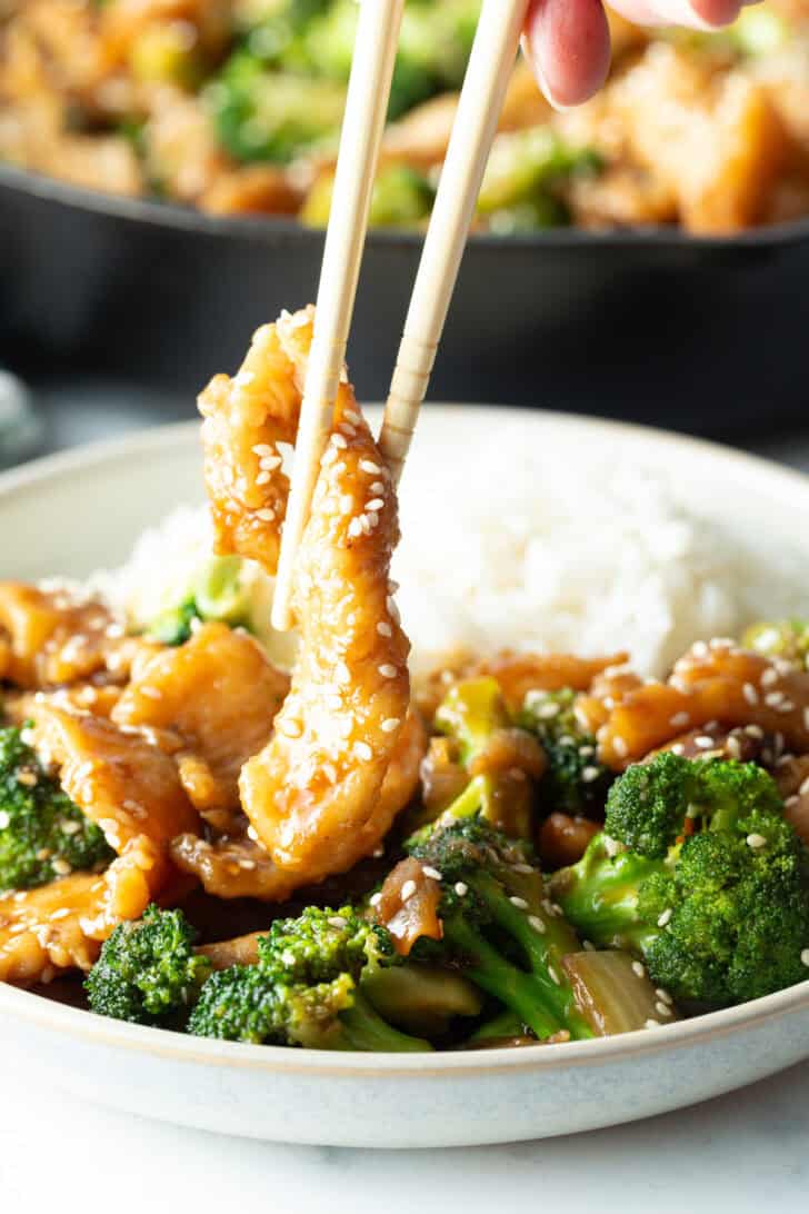 Pair of chopsticks grabbing a piece of chicken from the bowl of broccoli chicken.