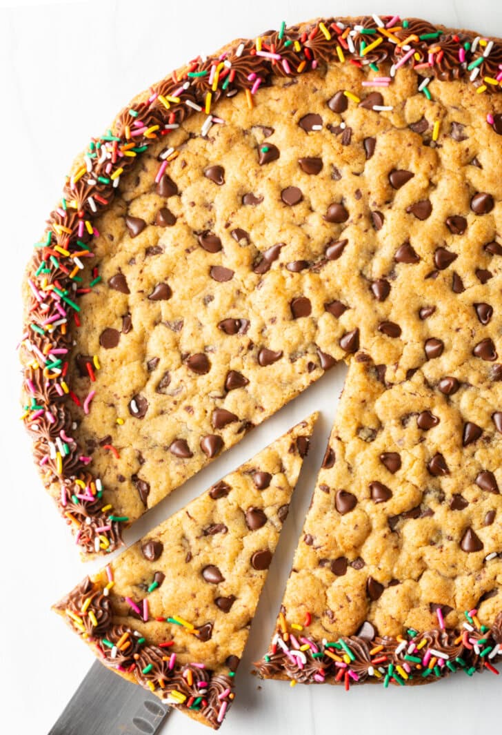 Large chocolate chip cookie cake. The outside edge is piped with chocolate frosting and covered with rainbow sprinkles. A cake cutter is taking a wedge slice from the cake.