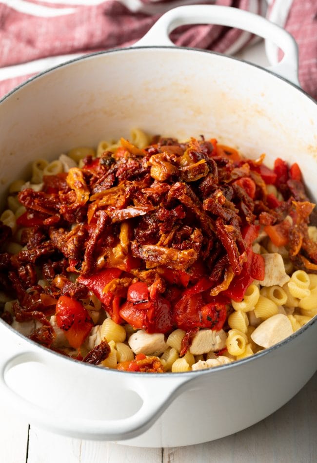 White cooking pot with chunks of cooked chicken and raw macaroni pasta noodles, topped with roasted red peppers and sun dried tomatoes.