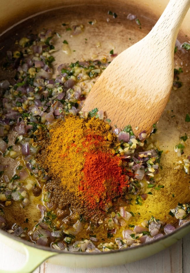 Spices and aromatics in a dutch oven being sautรฉed and stirred with a wooden spoon.