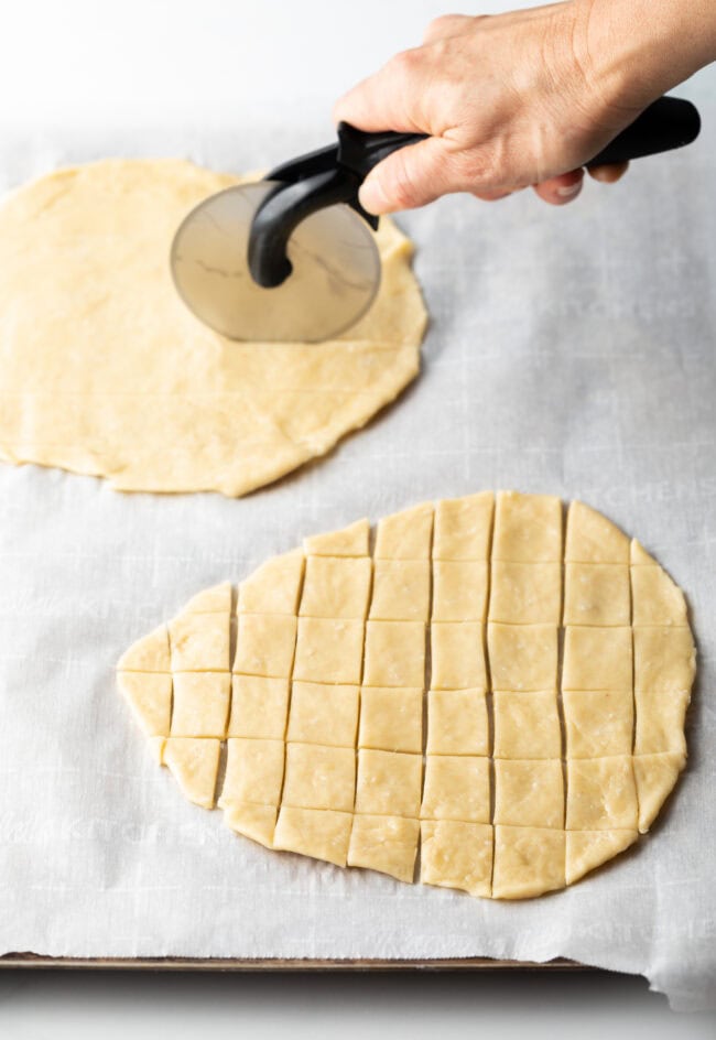 Hand with pizza cutter slicing two unbaked ovals of unleavened bread into tiny squares.