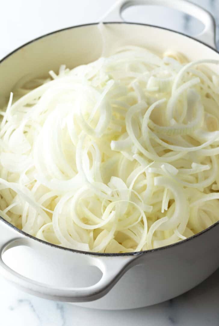 Large white cooking pot brimming with thinly sliced white onions.