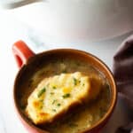 Large red mug with French onion soup, topped with a slice of baguette with melted cheese.