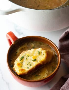 Large red mug with French onion soup, topped with a slice of baguette with melted cheese.
