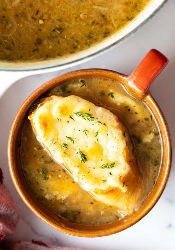 Top down view of a large red mug with French onion soup, topped with a slice of baguette with melted cheese.