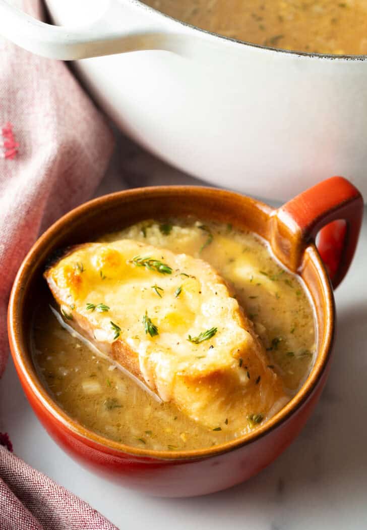 Large red mug with French onion soup, topped with a slice of baguette with melted cheese.