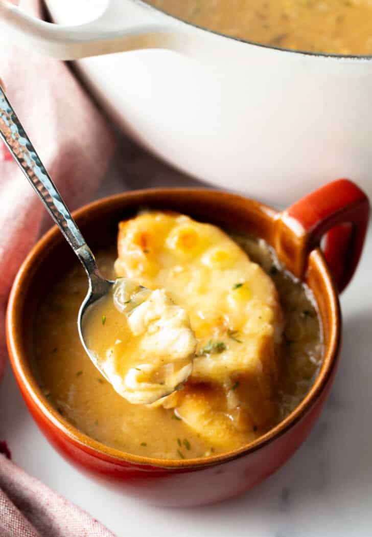 Large red mug with French onion soup, topped with a slice of baguette with melted cheese. A spoon is going in for a scoop.