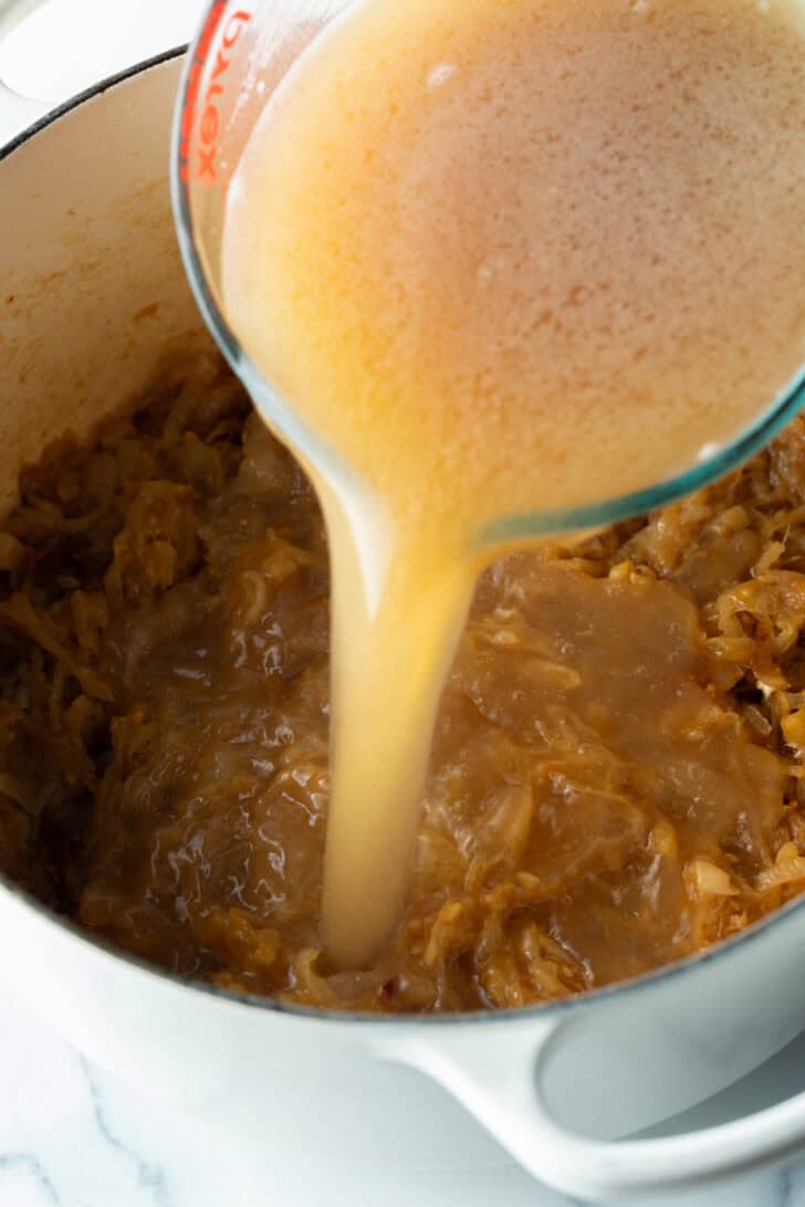 Pouring beef stock from a glass pitcher into the large pot with cooked onions.