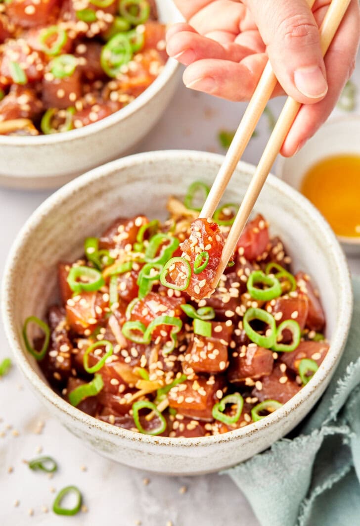 ahi-poke.jpg Hand holding a piece of raw tuna with chopsticks with more ahi poke in the background.
