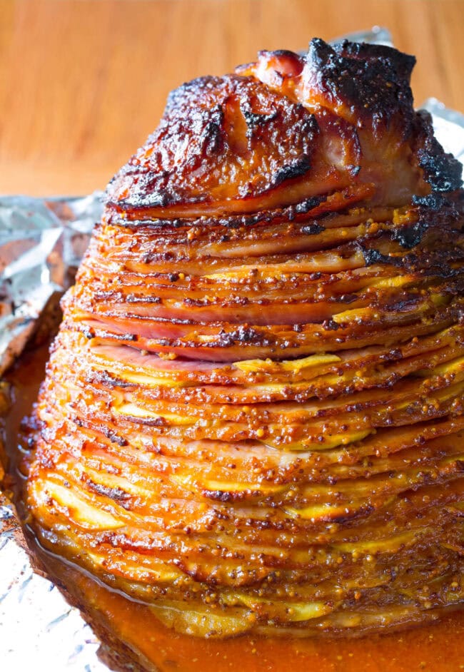 This Baked Ham Recipe is the perfect centerpiece to your holiday meal. Slathered with honey mustard ham glaze, stuffed with apples and baked to perfection! #ASpicyPerspective #Holiday # Easter #Christmas #Thanksgiving #Ham #BakedHam #BakedHamRecipe #HoneyMustard #Apples #HamGlaze Fully Cooked Apple Stuffed Ham with Mustard Glaze on the baking sheet.