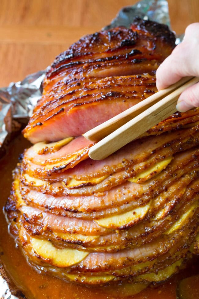 This Baked Ham Recipe is the perfect centerpiece to your holiday meal. Slathered with honey mustard ham glaze, stuffed with apples and baked to perfection! #ASpicyPerspective #Holiday # Easter #Christmas #Thanksgiving #Ham #BakedHam #BakedHamRecipe #HoneyMustard #Apples #HamGlaze Choosing the slices of ham with the apples with a pair of tongs.