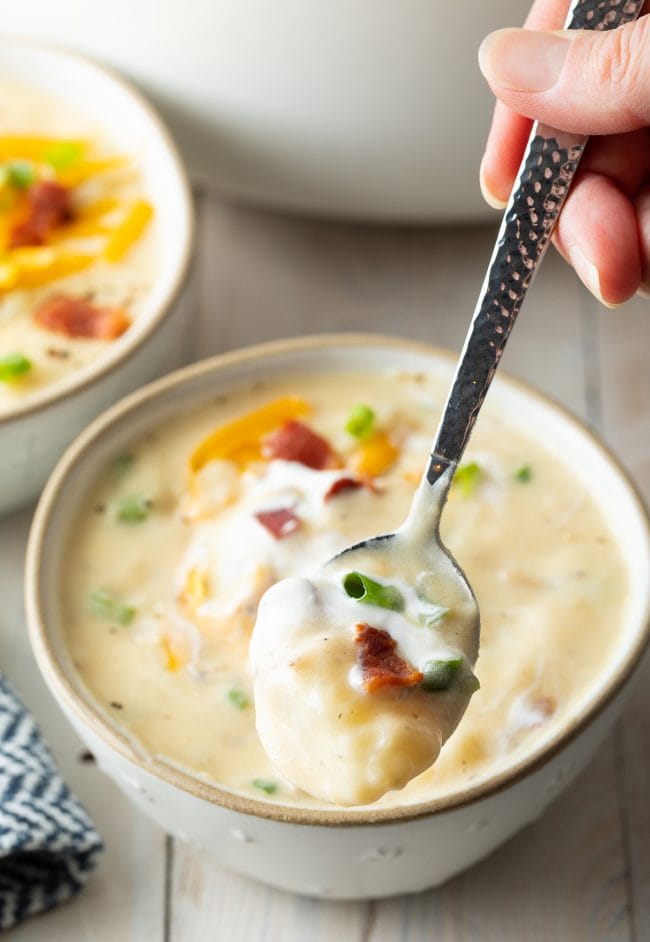 Spoon with potato soup on it and the bowl of soup in the background.