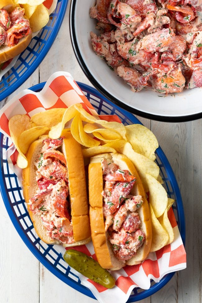 Overhead shot of lobster rolls in a serving basket with chips and more lobster in the background. 