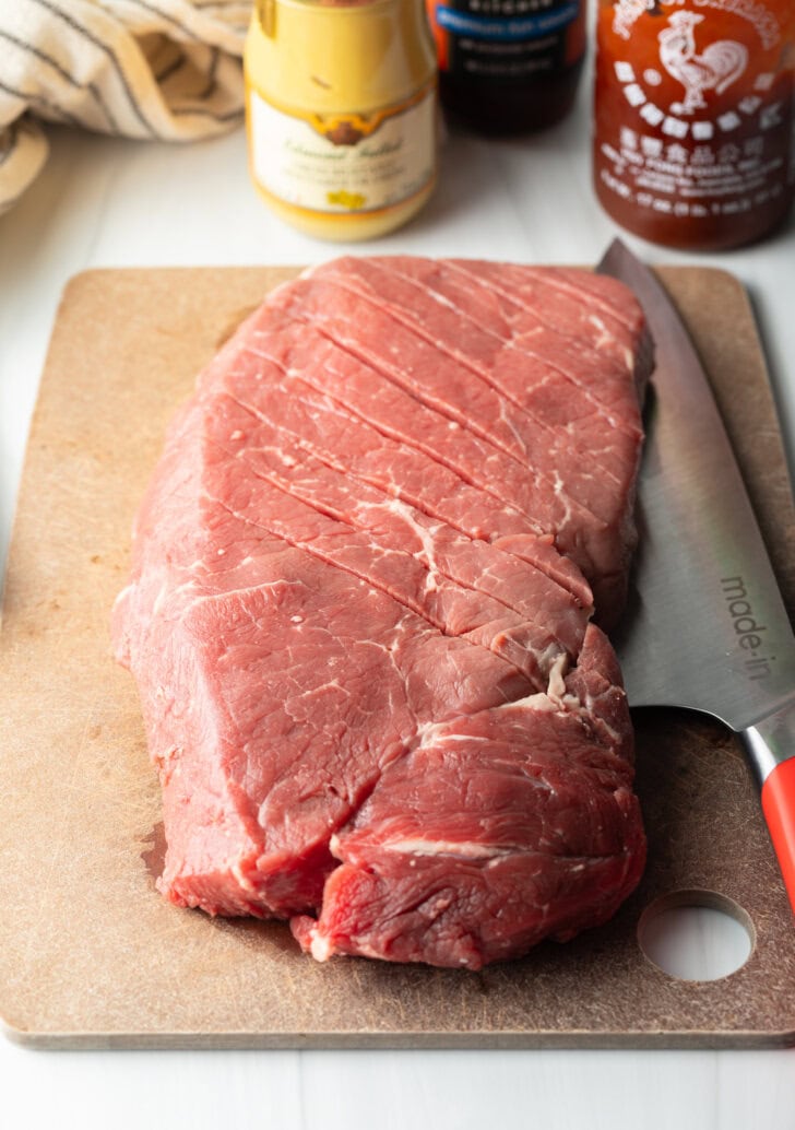 Knife scoring a large section of London broil on a cutting board with a criss-cross diagonal pattern.