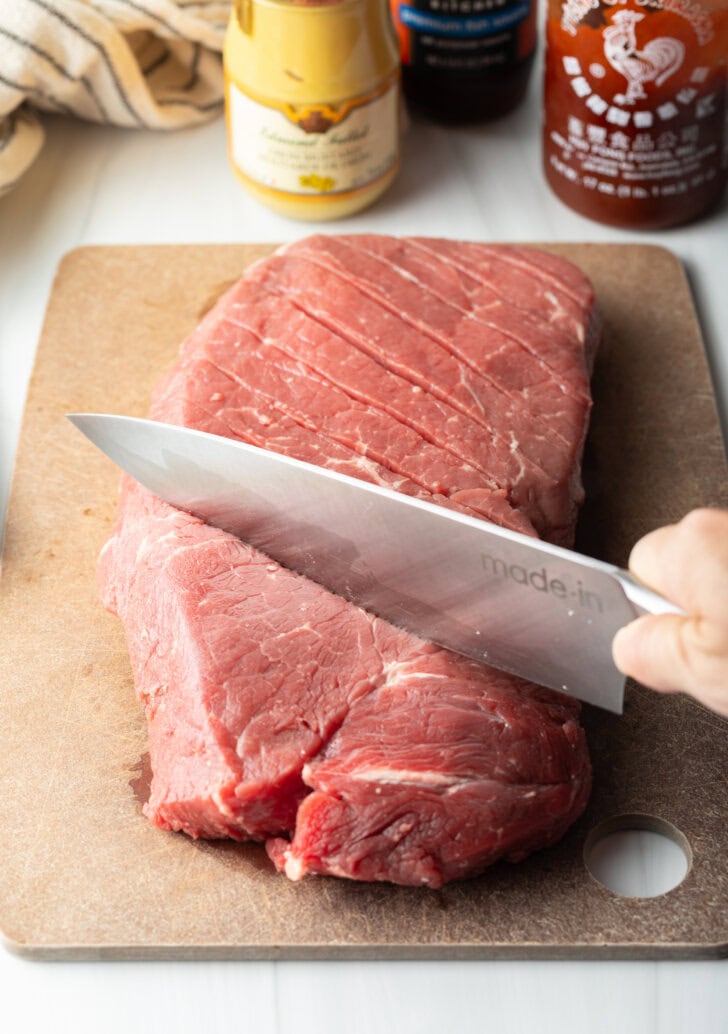 Knife scoring a large section of London broil on a cutting board with a criss-cross diagonal pattern.