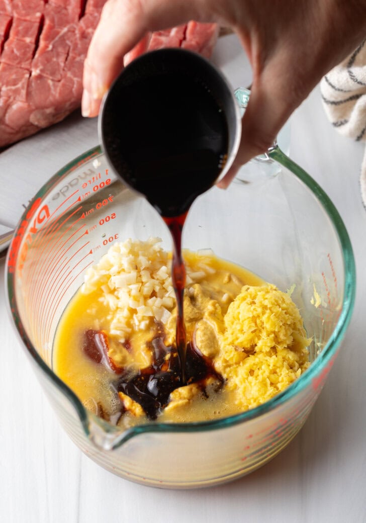 Hand adding dark brown sauce to glass measuring cup with marinade ingredients.