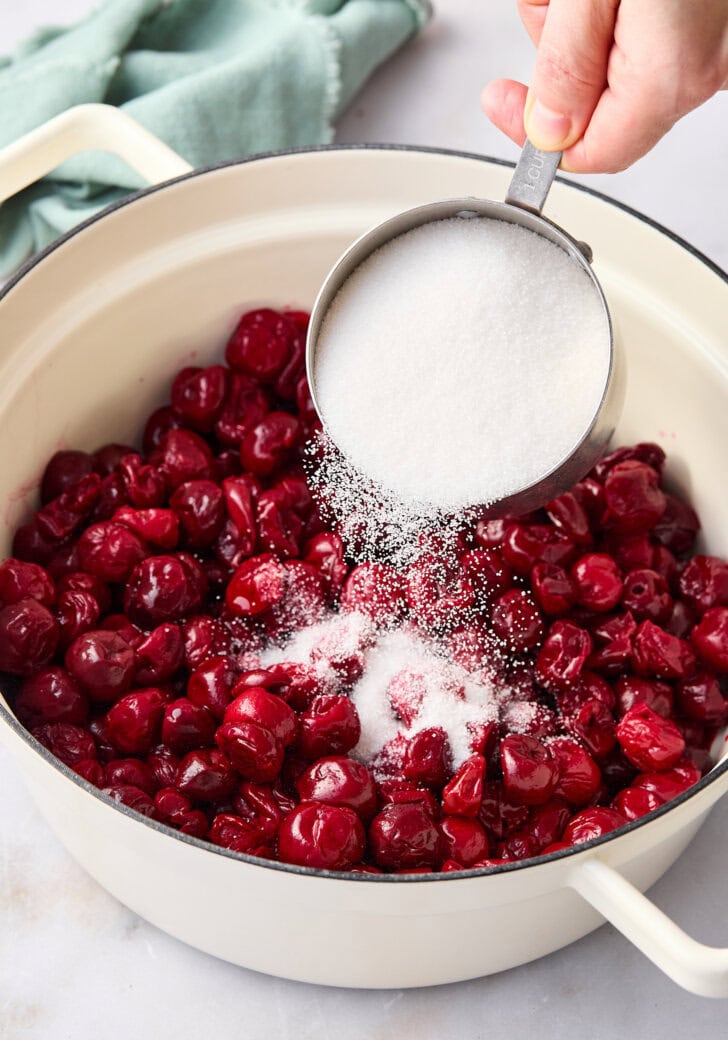 Hand pouring sugar into a pot filled with fresh cherries.