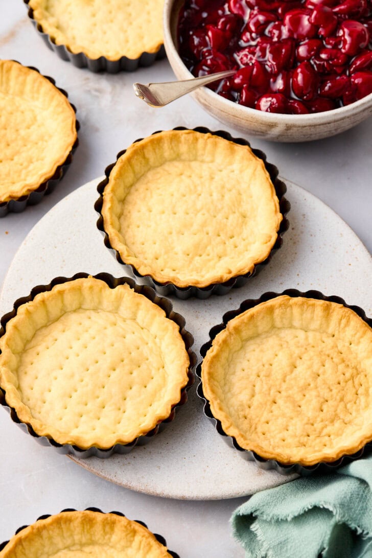 Baked cherry pie crusts in the tart pans next to a bowl of cherry pie filling.