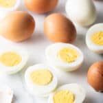 A dozen hard boiled eggs on a white marble backdrop. There are five egg halves, four brown eggs in the shell, and one white peeled egg.