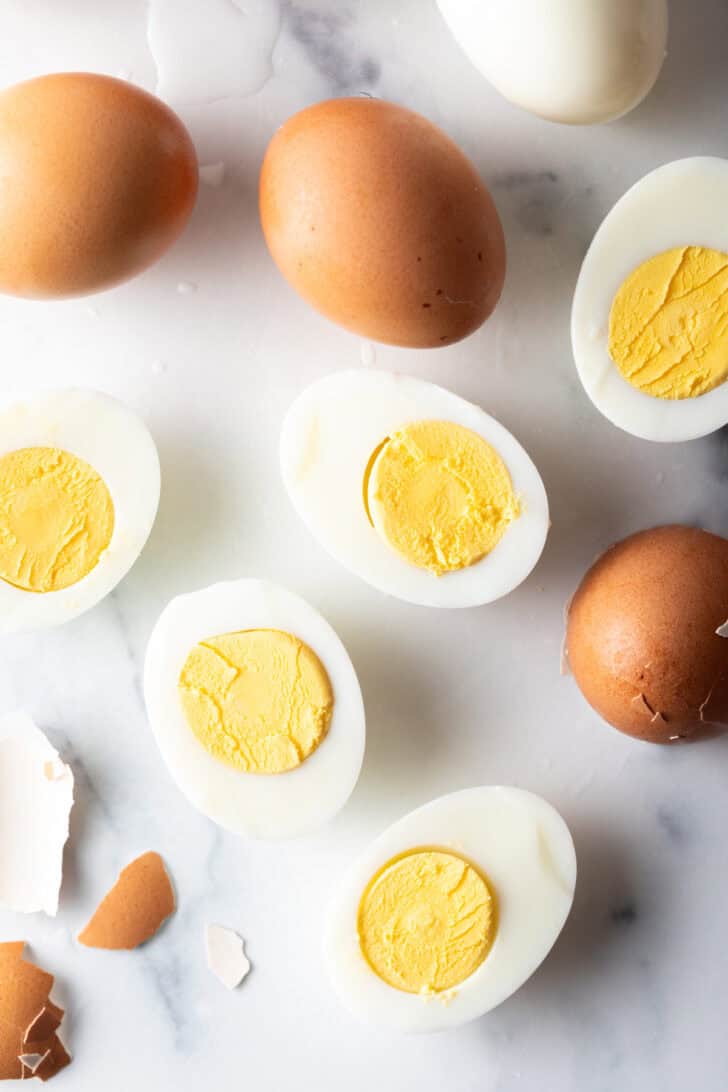 A dozen hard boiled eggs on a white marble backdrop. There are five egg halves, four brown eggs in the shell, and one white peeled egg.
