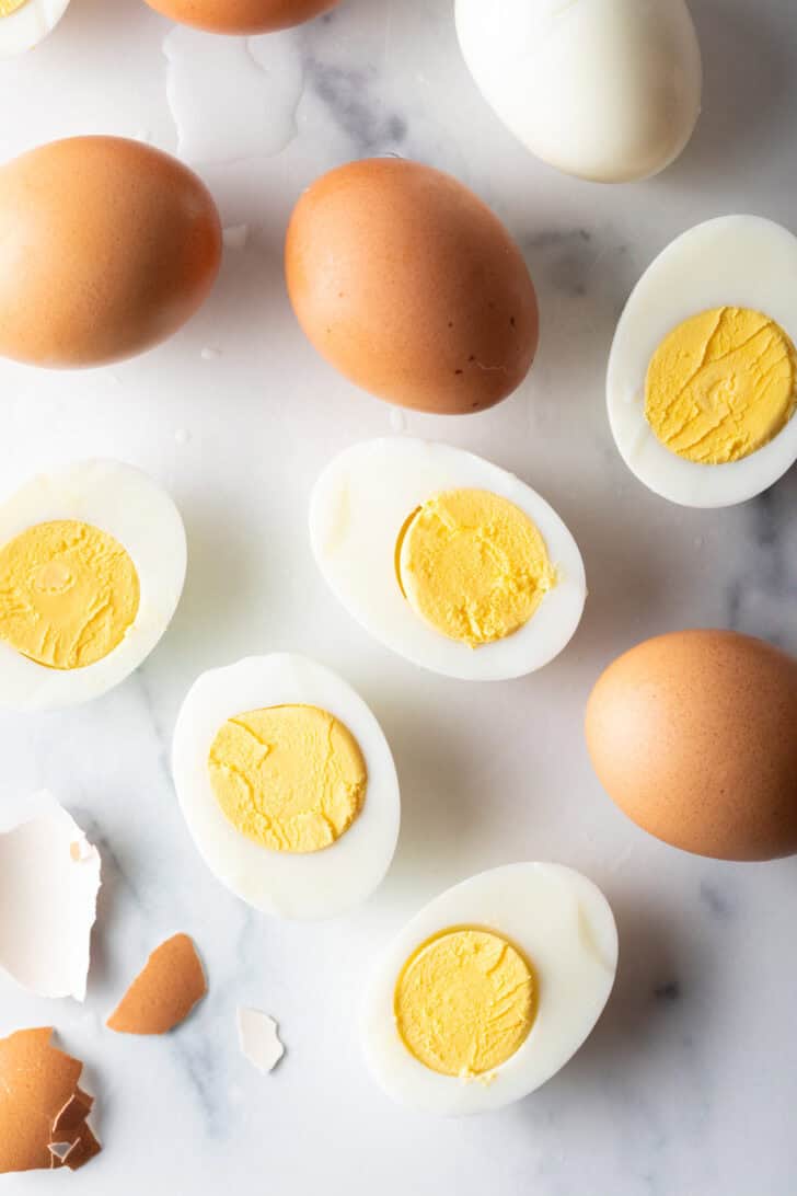 A dozen hard boiled eggs on a white marble backdrop. There are five egg halves, four brown eggs in the shell, and one white peeled egg.