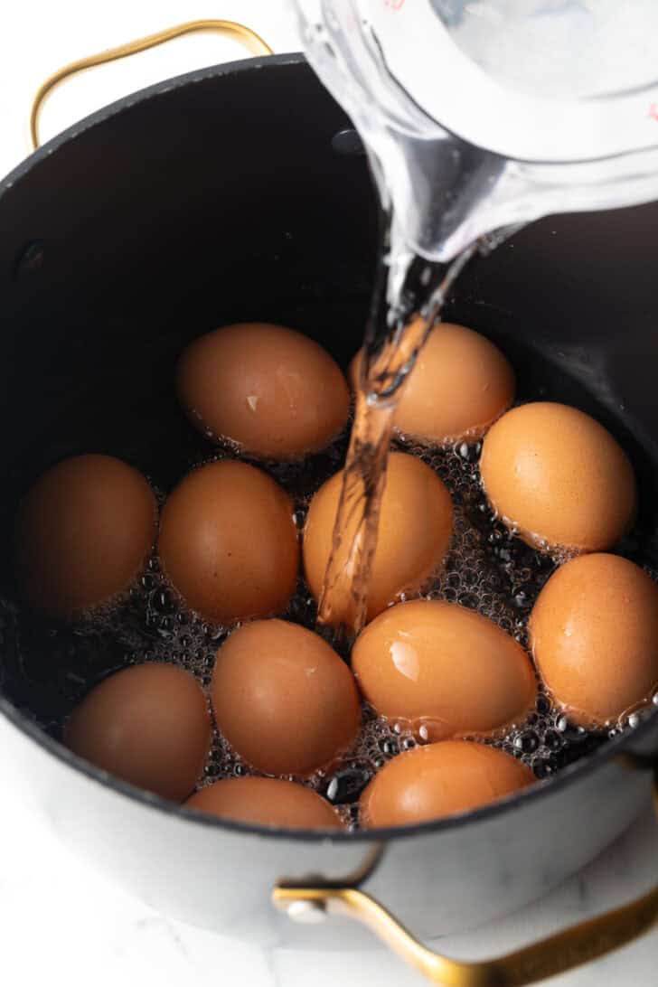 Pouring water from a glass measuring glass over a dozen brown eggs in a black cooking pot.
