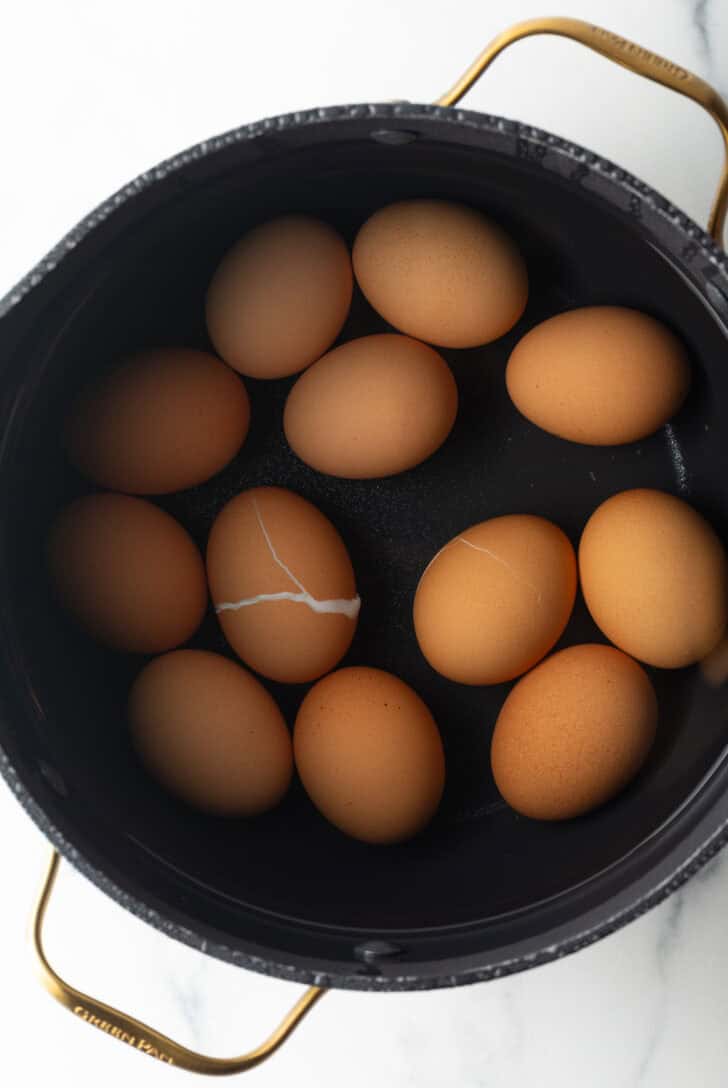 A dozen brown eggs covered in water in a large black pot.