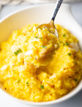 Metal spoon scooping a bite of yellow risotto milanese from a white bowl, showing to camera.