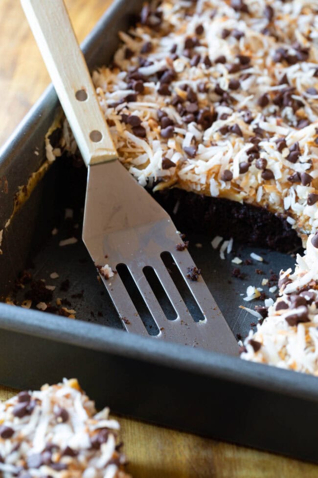 A tray of the baked and ready Samoas Texas sheet cake, which is topped with a creamy frosting, shredded coconut and mini chocolate chips. A large piece of the cake has been removed by a spatula.