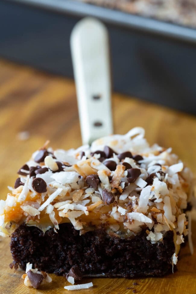 A slice of the Samoas Texas Sheet Cake sits atop a spatula. The cake is made up of a chocolate cake layer topped with caramel sauce, toasted coconut flakes and mini chocolate chips.