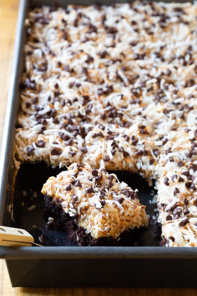 A tray of the baked and ready Samoas Texas sheet cake, which is topped with a creamy frosting, shredded coconut and mini chocolate chips. A large piece of the cake is being removed by a spatula.