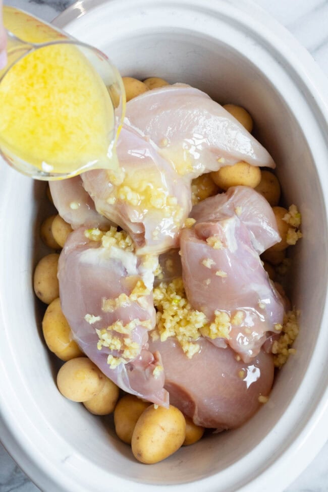 Raw chicken and potatoes in a slow cooker with a garlic butter mixture being poured over the top.