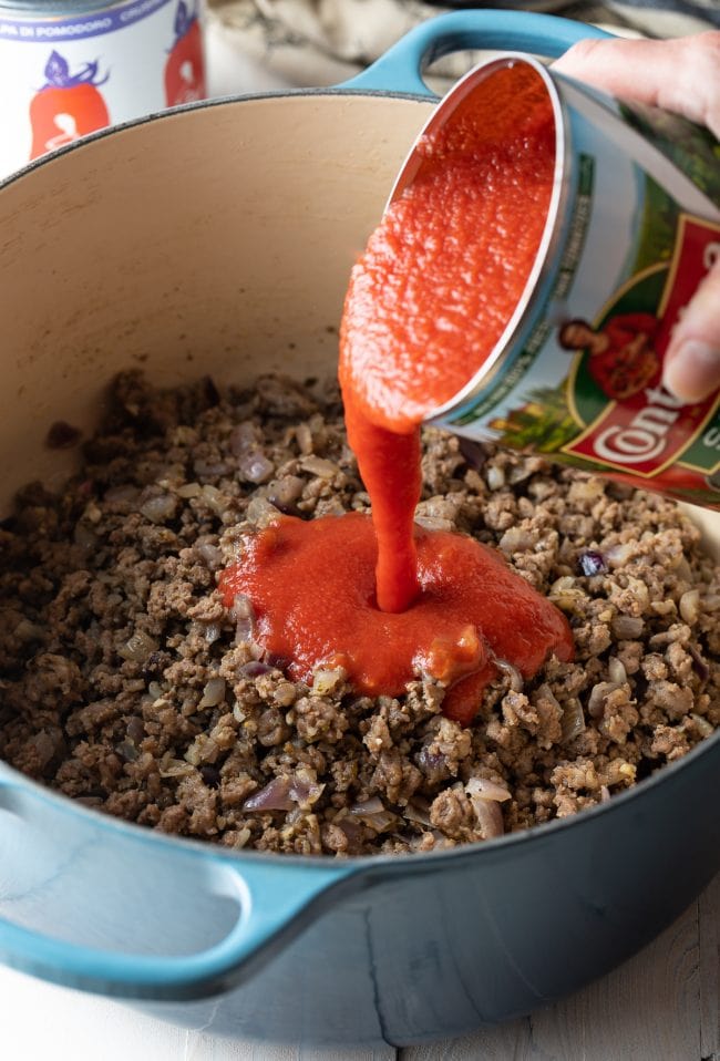 Hand pouring a can of tomato sauce into a pot with ground beef.